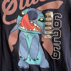Puede incluir: Una camiseta azul y blanca con un personaje de dibujos animados de Stitch que lleva una camisa azul y la lengua rosa que sobresale. La camiseta tiene la palabra "Stitch" en letras rosas y el n&uacute;mero "626" en letras blancas. La camiseta tiene una etiqueta blanca con la palabra "SMALL" repetida varias veces.
