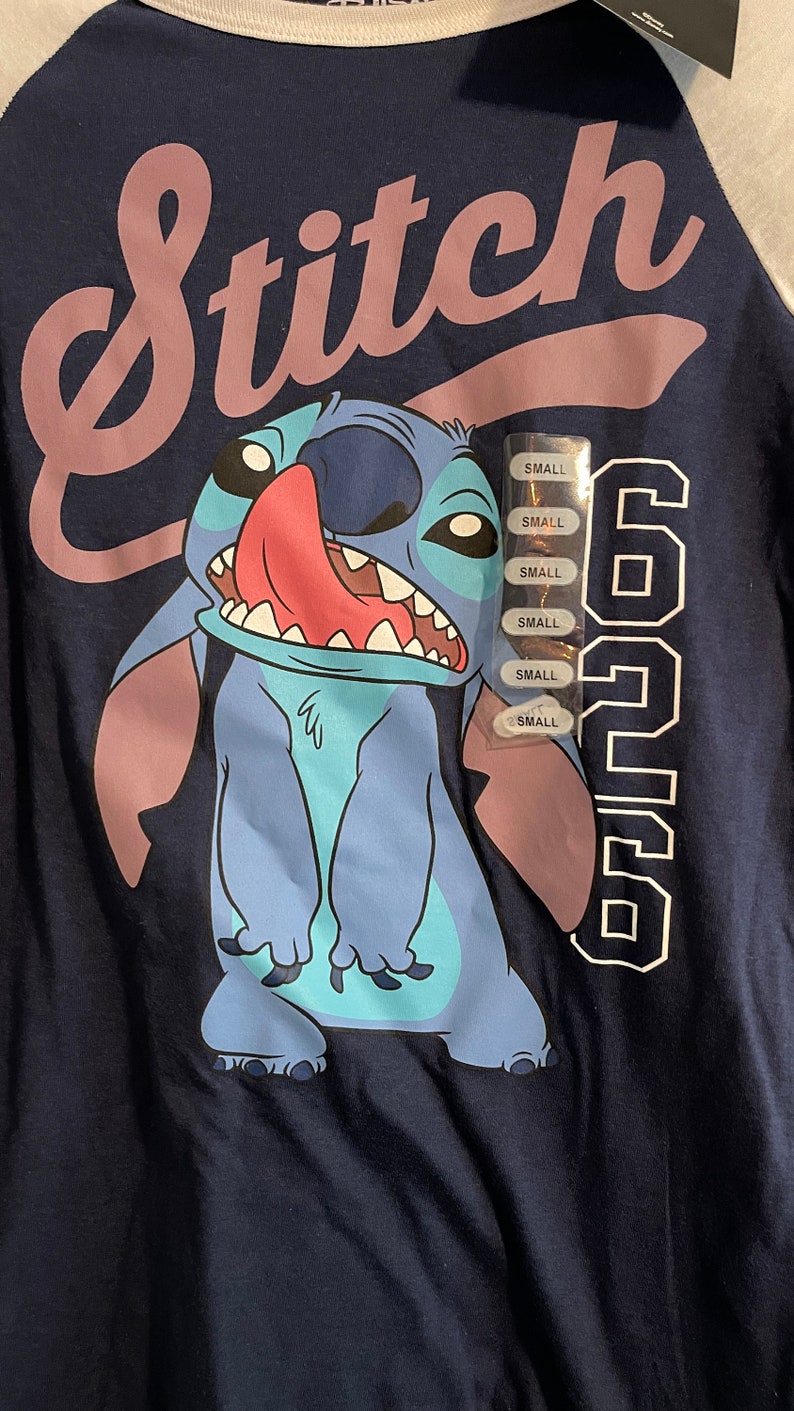 Puede incluir: Una camiseta azul y blanca con un personaje de dibujos animados de Stitch que lleva una camisa azul y la lengua rosa que sobresale. La camiseta tiene la palabra "Stitch" en letras rosas y el n&uacute;mero "626" en letras blancas. La camiseta tiene una etiqueta blanca con la palabra "SMALL" repetida varias veces.
