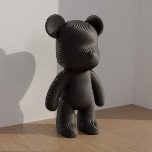 May include: A black, stylized bear figurine with a textured, layered design. The bear has rounded features, including ears, arms, and legs. It stands on a wooden surface, with a white wall in the background. The bear's shadow is visible on the wall.