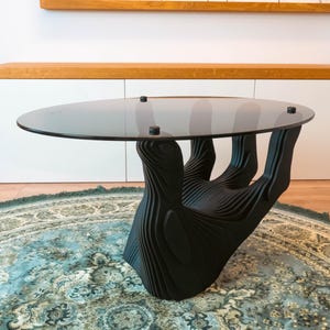 May include: A modern black coffee table with a unique hand-shaped base. The table features a round, clear glass top supported by a detailed, layered design that forms the shape of a hand. The table is set on a patterned rug.
