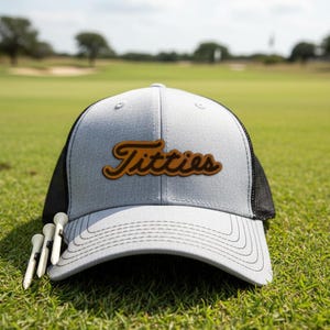 Pode incluir: Boné de caminhoneiro cinza e preto com a palavra "Titties" em escrita marrom. O boné está em um campo de golfe verde, com três tees de golfe brancos.