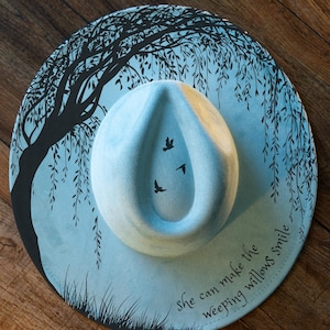 May include: A light blue fedora hat with a willow tree design and the text "She can make the weeping willows smile." The hat features three flying birds and a light blue crown.