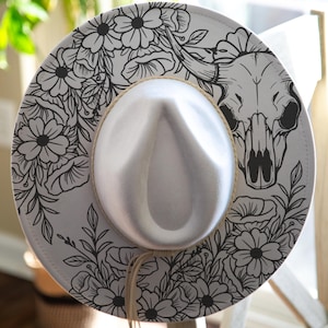 May include: A white fedora hat featuring a black line art illustration of a bull skull surrounded by various floral patterns on the brim. The hat includes a light tan cord band wrapped around the crown with two thin hanging cord ends.