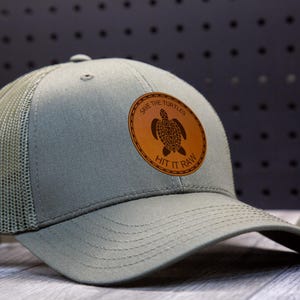 Könnte beinhalten: Eine Trucker-Cap in Salbeigrün mit Mesh-Rückseite und gebogenem Schirm. Ein kreisförmiger Lederaufnäher zeigt ein Schildkröten-Design und den Text "Save the Turtles" und "Hit it Raw".