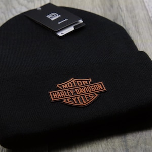 May include: Black knit beanie featuring a Harley-Davidson logo patch in brown and orange. The beanie has a folded cuff and a tag attached. The logo includes the words "MOTOR HARLEY-DAVIDSON CYCLES".