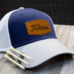 May include: A navy blue and grey baseball cap with a white mesh back. The cap has a brown leather patch with the word "Titties". Three wooden golf tees are placed in front. The cap is a popular style.