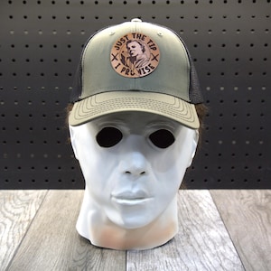 May include: An olive green and black trucker hat with a patch saying "JUST THE TIP I PROMISE". The hat has a mesh back and a curved peak. The patch features a vintage style image.