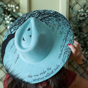 May include: A light blue wide-brimmed hat featuring a black hand-painted design of a weeping willow tree, three flying birds, and the text "she can make the weeping willows smile" written in a cursive script along the brim edge.
