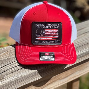 May include: Red and white trucker hat with a red brim and crown, white mesh back, and a rectangular patch. The patch features a distressed American flag design and the text "WHEN TYRANNY BECOMES LAW REBELLION BECOMES DUTY."