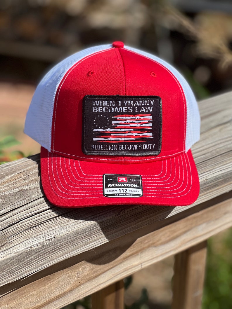 May include: Red and white trucker hat with a red brim and crown, white mesh back, and a rectangular patch. The patch features a distressed American flag design and the text "WHEN TYRANNY BECOMES LAW REBELLION BECOMES DUTY."