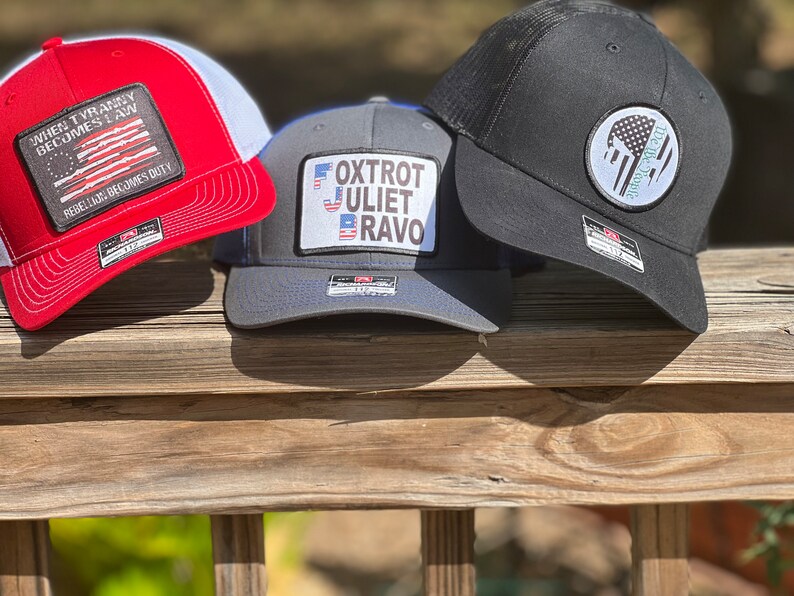 May include: Three baseball caps with different designs. One is red and white with the text "WHEN TYRANNY BECOMES LAW REBELLION BECOMES DUTY". Another is gray with the text "FOXTROT JULIET BRAVO". The third is black with a white patch.