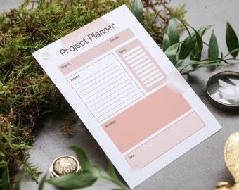 Puede incluir: Un planificador de proyectos blanco con acentos rosados. El planificador tiene secciones para el proyecto, la actividad, la fecha límite, los objetivos, la estrategia y las notas.