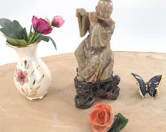 May include: A green jade sculpture of a person playing a flute, sitting on a wooden base. The sculpture is on a wooden surface with a vase of pink flowers, a peach-colored rose, and a blue and gold butterfly.