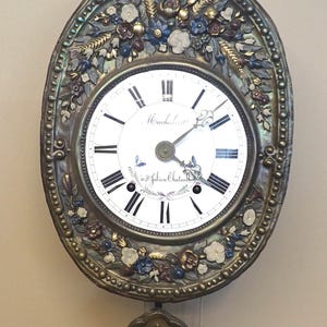 Könnte beinhalten: Eine ovale antike Uhr mit weißem Zifferblatt und schwarzen römischen Ziffern. Die Uhr ist in einem verzierten Metallrahmen mit floralen und Weizenähren-Designs in Blau, Gold und Weiß eingefasst. Das Zifferblatt zeigt "Machalasse" und "à St Julien Chatenay".