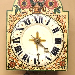 Puede incluir: Un reloj de pared vintage con una esfera crema y números romanos negros. El reloj está adornado con un diseño floral con rosas rojas y hojas verdes. Las manecillas del reloj son negras y doradas, y tiene dos pesas colgantes.