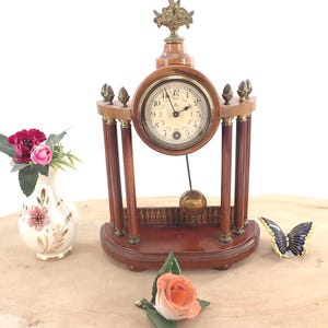May include: A wooden mantel clock with a brass pendulum and a decorative floral design on top. The clock face has Roman numerals and a gold trim.