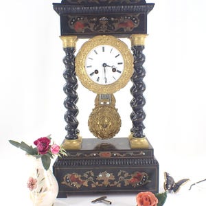 May include: An antique black mantel clock with gold accents and a white clock face. The clock features a decorative pendulum and is flanked by a vase of flowers, a rose, and a butterfly figurine. The clock's height is approximately 24 inches.