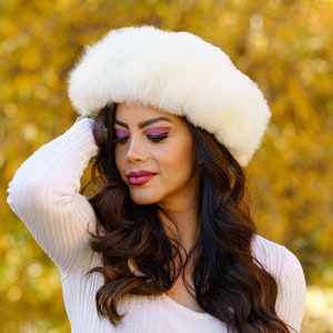 May include: A woman wearing a white, fluffy hat and a light pink, ribbed long-sleeve shirt. She is also wearing blue jeans. The background is a blurred view of fall foliage.