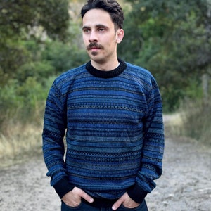May include: A man wearing a blue and black striped jumper with a black collar and blue jeans. He is standing on a dirt path with green trees and hills in the background.