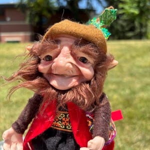 Könnte beinhalten: Eine handgefertigte Zwergenpuppe mit detailliertem Gesicht, braunem Haar und einem braunen Hut. Der Zwerg trägt eine rote Weste über einem gemusterten Hemd, eine schwarze Hose und einen kleinen Rucksack. Die Puppe ist im Freien vor einem verschwommenen grünen Hintergrund posiert.
