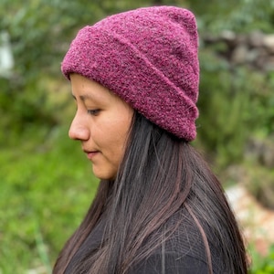 May include: A close-up of a magenta-colored beanie hat. The hat is made of a textured knit material and has a folded brim. The hat is worn by a person with long, dark hair and a black shirt. The background is blurred with green foliage.