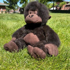 Puede incluir: Un peluche de gorila con pelaje marrón oscuro, una cara marrón y grandes pies marrones. El gorila está sentado sobre hierba verde en un entorno soleado al aire libre. El juguete tiene una boca roja y una apariencia suave y tierna.