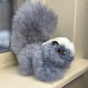 Puede incluir: Un juguete de peluche gris y blanco con forma de mofeta. El juguete tiene una cola larga y tupida y una raya negra en la espalda.