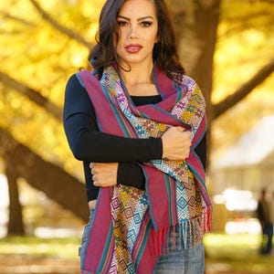 Puede incluir: Una mujer con una camisa negra de manga larga y pantalones vaqueros azules con una bufanda de estampado colorido drapeada alrededor de su cuello. La bufanda tiene flecos y está hecha de una tela suave y tejida.