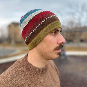 May include: A striped knit beanie with a brown, green, red, white, and blue color scheme. The beanie has a row of silver beads around the brim.