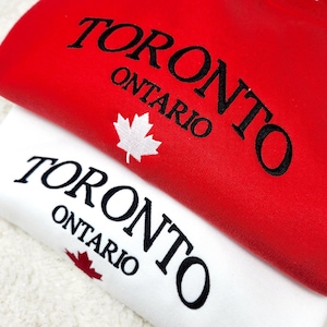 May include: Two sweatshirts, one red and one white, are stacked. The red sweatshirt has "TORONTO ONTARIO" embroidered in black, with a white maple leaf. The white sweatshirt has the same text and a red maple leaf.