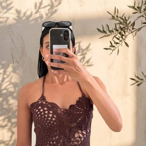 May include: A brown crochet halter top with a unique openwork design. The top features a scalloped edge and a halter neckline. The model is wearing sunglasses on their head and holding a phone in front of their face. The background is a neutral-toned wall.