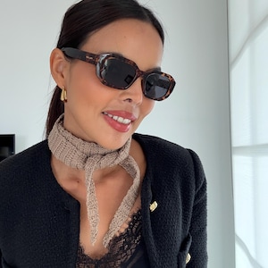 May include: Tortoiseshell sunglasses with dark lenses. A beige knitted scarf is worn with a black jacket with gold buttons. The jacket is over a black top with lace detailing.