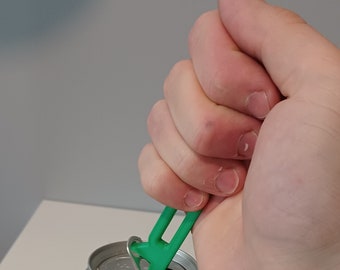 May include: A green can opener is used to lift the tab on a silver aluminum can. The can opener is held in a hand. The background is a plain, light gray wall.