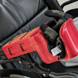 May include: A red and orange 3D-printed object containing black and green work gloves. The object, with a rectangular design and cut-out sections, rests on a black chair. The gloves are visible inside.