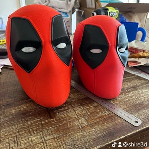 May include: Two red and black Deadpool masks with a white mesh eye covering. The masks are sitting on a wooden table.