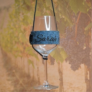 Könnte beinhalten: Ein blauer Filz-Weinglas-Anhänger mit dem Namen "Sarah" bestickt. Der Anhänger hängt an einer schwarzen Schnur und ist an einem klaren Weinglas befestigt.