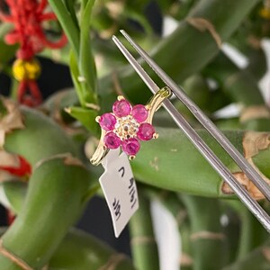 May include: A gold ring featuring a flower-shaped cluster of six round, vibrant pink gemstones surrounding a small, central cluster of tiny clear stones. A white rectangular jewelry tag with black printed text is attached to the gold band.