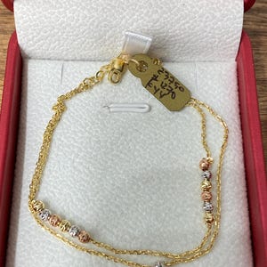 May include: A delicate gold chain bracelet featuring two strands and small, alternating textured beads in yellow, white, and rose gold tones. The bracelet includes a lobster claw clasp and a small tan paper tag with the handwritten text 23750 #1270.