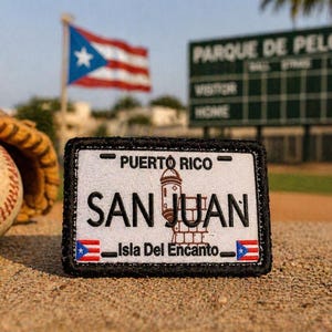 May include: A white license plate patch with black trim, featuring "SAN JUAN" in bold letters, the Puerto Rico flag, and the text "Isla Del Encanto." A baseball and glove are in the background, with the Puerto Rican flag waving.