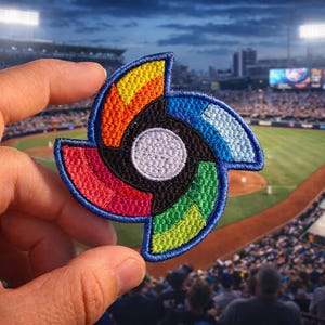 May include: Embroidered patch with a colorful, abstract design featuring red, orange, yellow, green, and blue segments. The patch has a white circle in the center and a blue border. The background shows a baseball stadium filled with spectators.