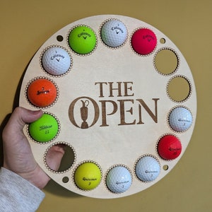 May include: A circular wooden golf ball display with the text "THE OPEN" and the Open Championship logo. The display holds golf balls of various colors, including white, green, orange, and red. The display is held by a person.