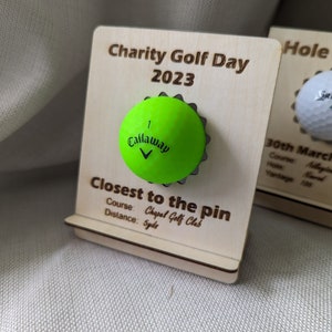 May include: A wooden plaque with the text "Charity Golf Day 2023" and "Closest to the pin" with a green golf ball attached. The text below the golf ball reads "Course: Chapel Golf Club" and "Distance: Syds".