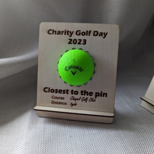 May include: A wooden plaque with the text "Charity Golf Day 2023" and "Closest to the pin". The plaque also includes the text "Course: Chapel Golf Club" and "Distance: Syde". A green golf ball with the Callaway logo is displayed on the plaque.