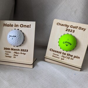 May include: Two wooden plaques with golf balls attached. The plaque on the left says "Hole in One!" and has a white golf ball with the text "SRIXON 4". The plaque on the right says "Charity Golf Day 2023" and has a green golf ball with the text "Callaway 1".