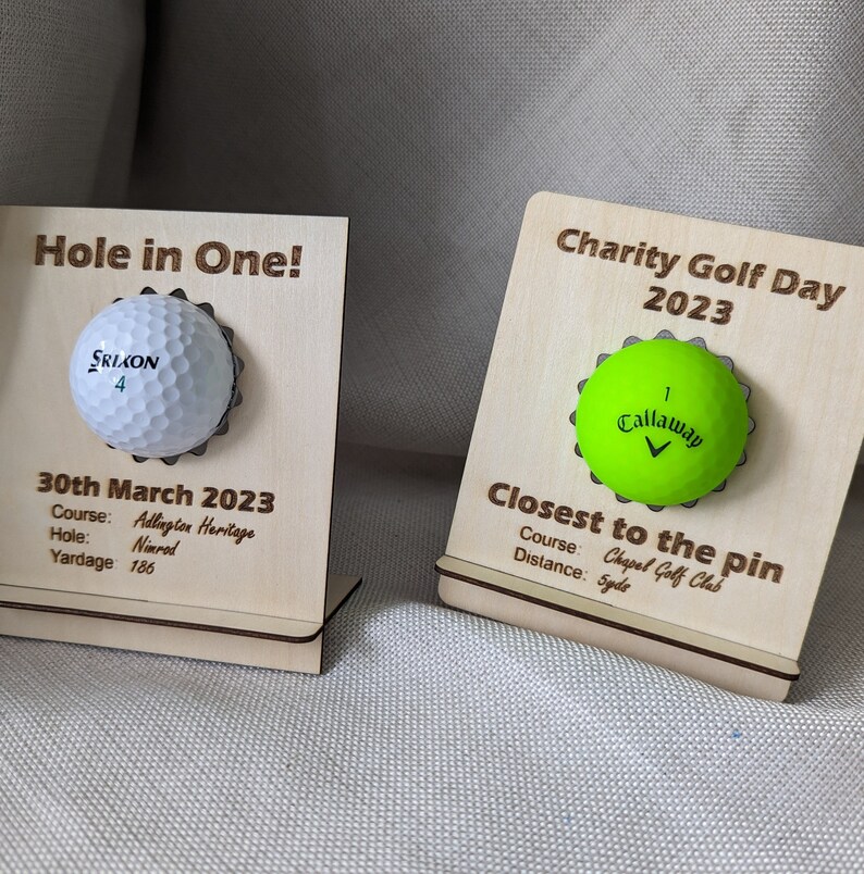 May include: Two wooden plaques with golf balls attached. The plaque on the left says "Hole in One!" and has a white golf ball with the text "SRIXON 4". The plaque on the right says "Charity Golf Day 2023" and has a green golf ball with the text "Callaway 1".