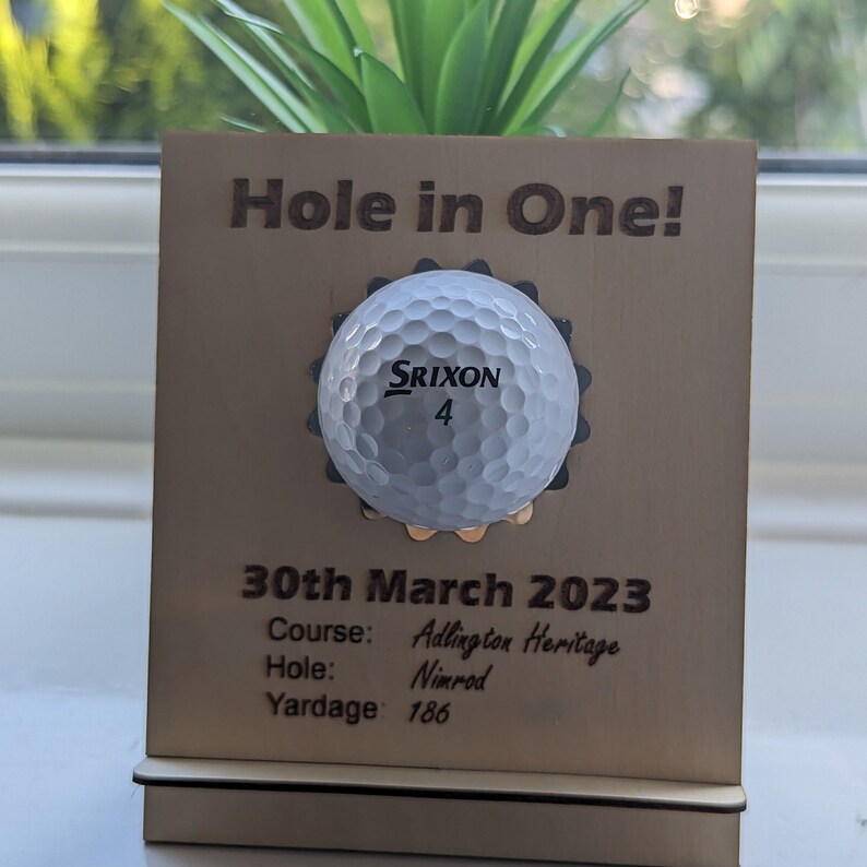 May include: A wooden plaque with a golf ball embedded in the center. The plaque reads "Hole in One!" and includes the date "30th March 2023", the course "Adlington Heritage", the hole "Nimrod", and the yardage "186".