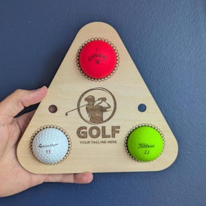 May include: A triangular light-colored wooden golf ball display rack featuring three circular cutouts holding a red Callaway ball, a white TaylorMade ball, and a lime green Titleist ball. The center is engraved with a golfer icon and the text GOLF YOUR TAGLINE HERE.