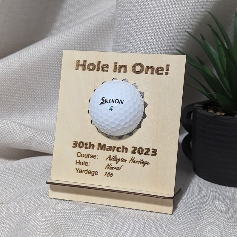 May include: A wooden plaque with a golf ball embedded in it. The plaque reads "Hole in One!" and includes the date "30th March 2023", the course "Adlington Heritage", the hole "Nimrod", and the yardage "186".