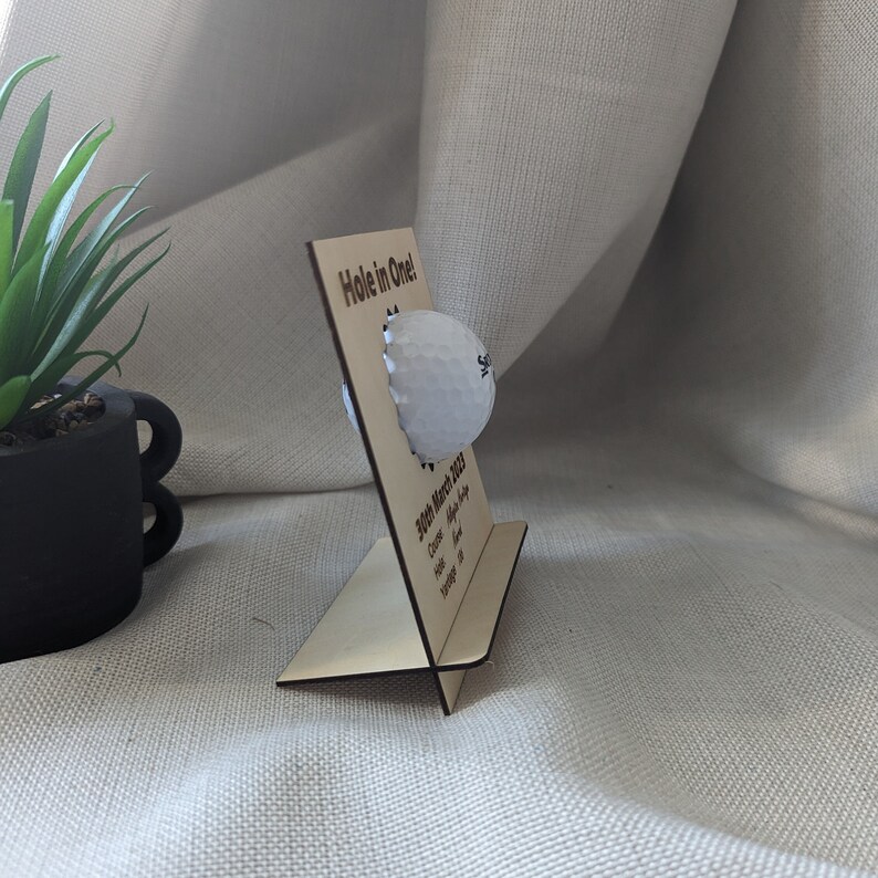 May include: A wooden plaque with the text "Hole in One!" and a golf ball attached. The plaque also includes the date "20th March 2023" and the words "Course, Hole, Alpha, Score, Handicap, Yardage".