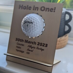 May include: A wooden plaque with a golf ball embedded in it. The plaque reads "Hole in One!" and includes the date "30th March 2023", the course "Addington Heritage", the hole "Nimrod", and the yardage "186".